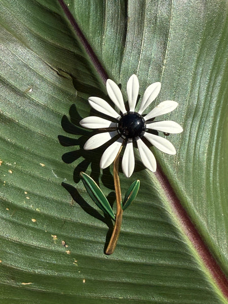 Vintage white enamel daisy brooch with black centre green leaves and gold tone stem displayed on a green leaf