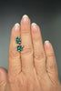A close-up of a hand wearing a pair of small cluster stud earrings with turquoise-like green stones on a dark gray background.
