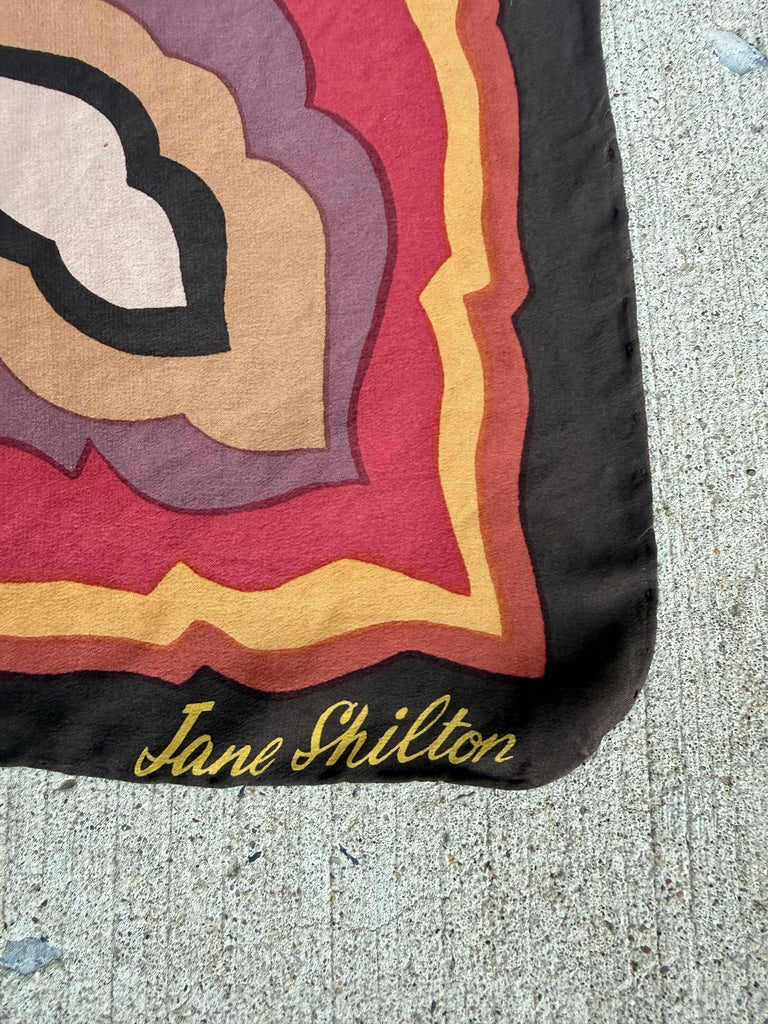 A close-up of a vintage Jane Shilton scarf laid flat. The scarf features a retro concentric floral pattern in shades of red, yellow, purple, beige, and black, with 'Jane Shilton' embroidered in gold script at the bottom corner.