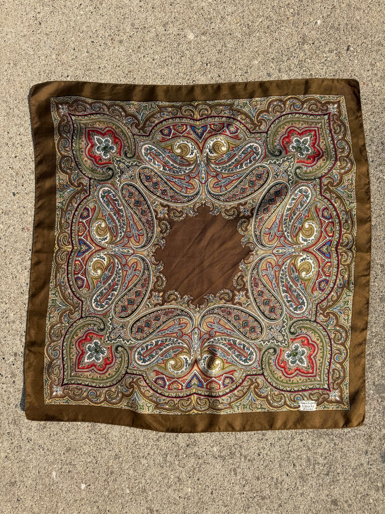 A square silk scarf with an intricate paisley pattern, laid flat. The design features a central brown medallion surrounded by detailed paisley motifs in red, gold, blue, and green, all framed by a solid brown border.