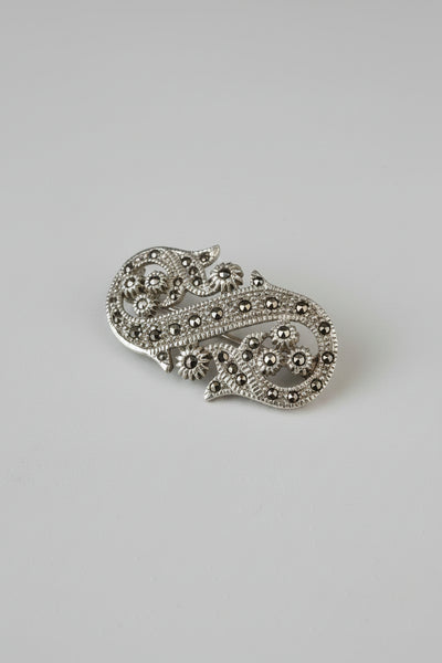 An ornate gold-toned filigree brooch with small dark gemstone accents, photographed on a light beige surface with a soft shadow against a pale gray background.