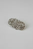 An ornate gold-toned filigree brooch with small dark gemstone accents, photographed on a light beige surface with a soft shadow against a pale gray background.