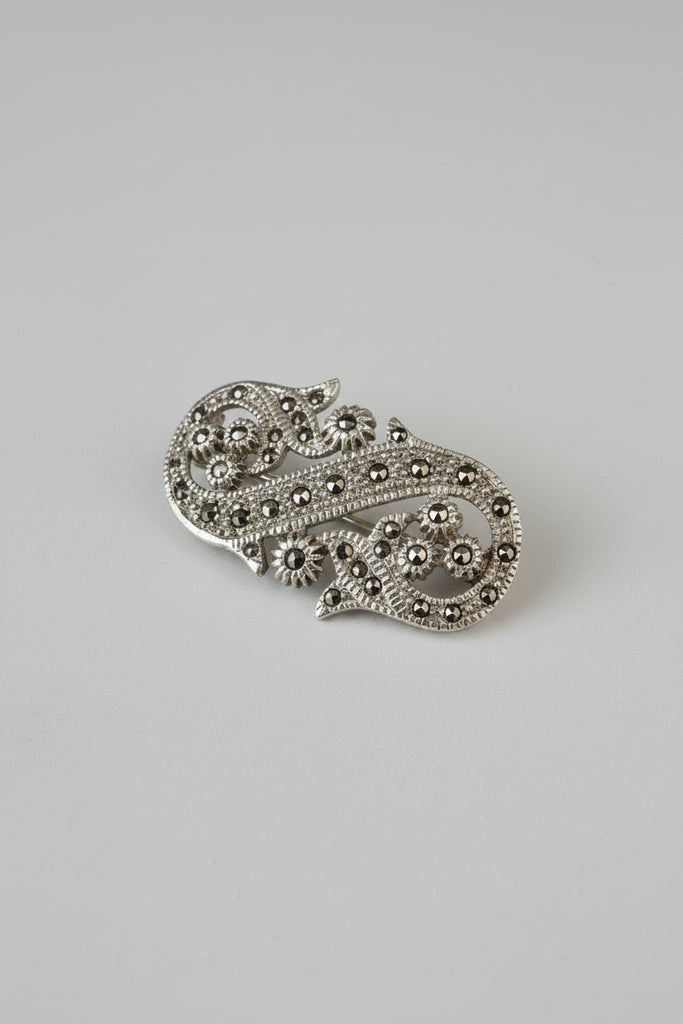 An ornate gold-toned filigree brooch with small dark gemstone accents, photographed on a light beige surface with a soft shadow against a pale gray background.