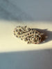 A vintage-style filigree brooch with intricate floral and scrollwork details, set in a gold-tone metal with small dark stones, resting on a light beige surface under direct sunlight.
