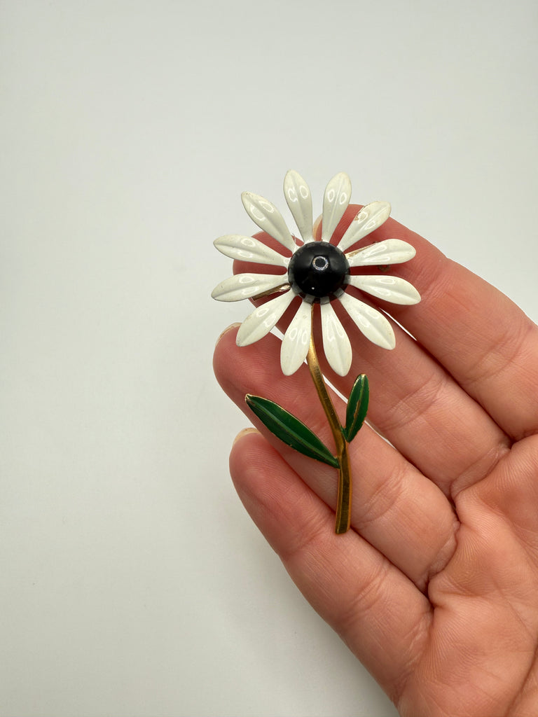 White enamel daisy brooch with dark green leaves and slender gold tone stem shown in hand for scale
