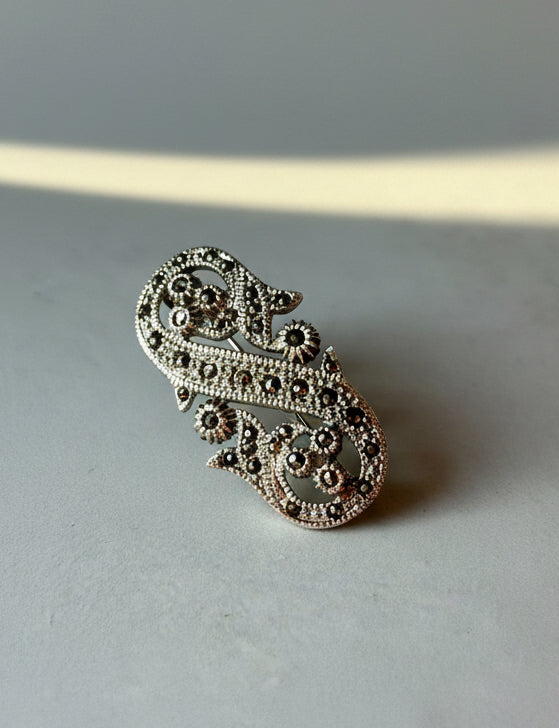 An ornate silver-toned paisley brooch embellished with small dark stones, resting on a plain gray surface. Sterling Silver 