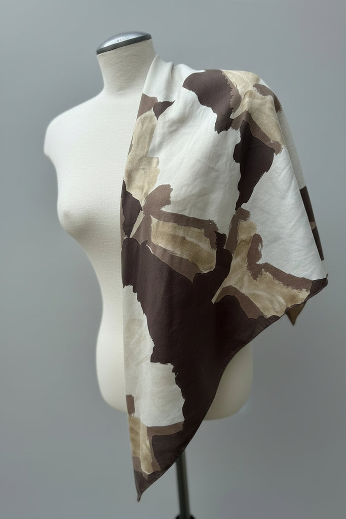 A lightweight scarf with a brown, beige, and off-white abstract camouflage pattern draped over a cream-colored dress form mannequin, set against a white garage door background.