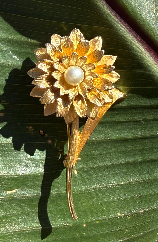 Vintage gold tone flower brooch with layered textured petals and faux pearl centre displayed on a green leaf