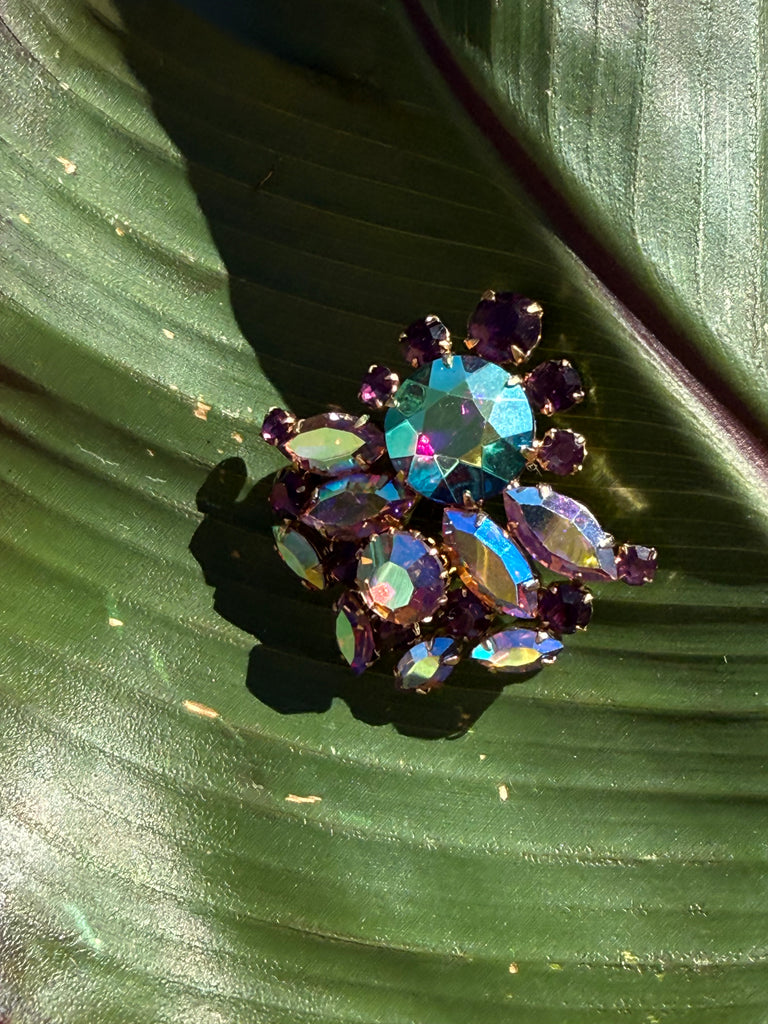 Vintage aurora borealis rhinestone brooch with iridescent multicolour stones and purple accents in a gold tone setting on green leaf