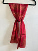 A red and gold patterned scarf tied in a knot and draped down a black hanger against a textured white wall.