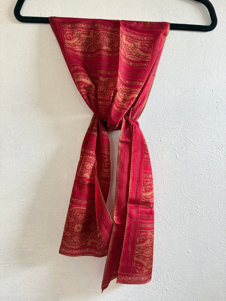 A red and gold patterned scarf tied in a knot and draped down a black hanger against a textured white wall.