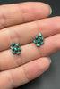 A close-up of a person's hand wearing a pair of small cluster stud earrings with turquoise-colored oval stones set in dark metal.
