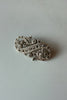 An ornate silver-toned filigree brooch with floral and scroll motifs, photographed on a neutral gray surface with a warm light reflection at the top.