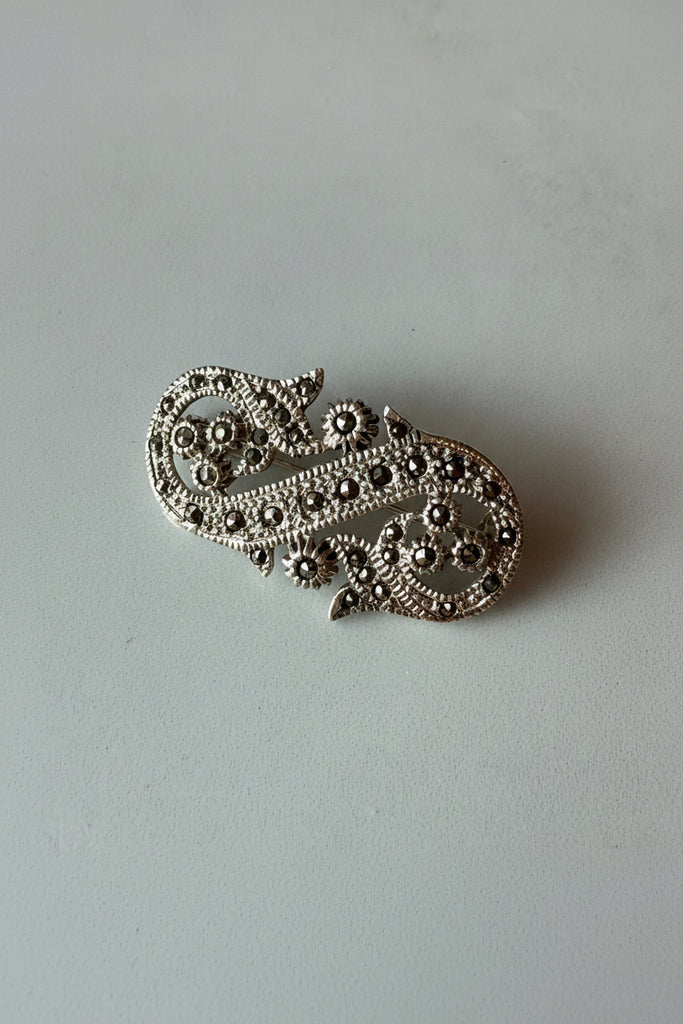 An ornate silver-toned filigree brooch with floral and scroll motifs, photographed on a neutral gray surface with a warm light reflection at the top.