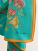 A close-up of this turquoise silk scarf with a floral print in pink, peach, and yellow, finished with a bright orange border. The signature 'Jain Thompson' is printed in black script near the bottom.