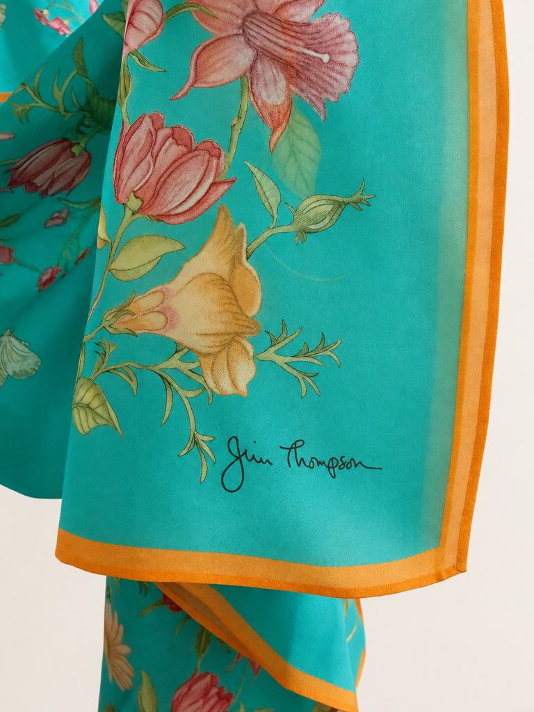 A close-up of this turquoise silk scarf with a floral print in pink, peach, and yellow, finished with a bright orange border. The signature 'Jain Thompson' is printed in black script near the bottom.