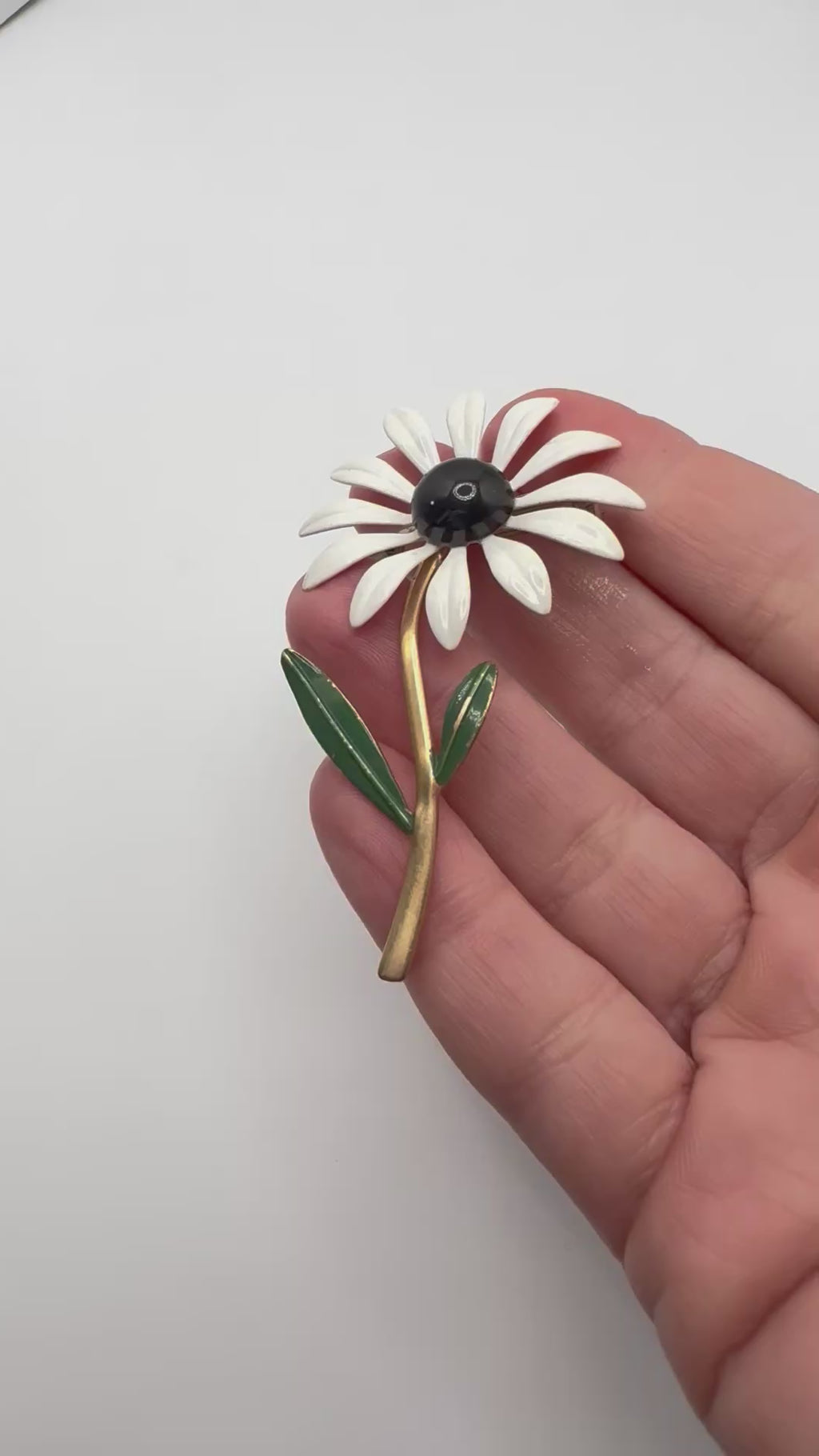 Vintage enamel daisy flower brooch shown in hand with white petals black centre and green leaves